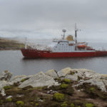 Large red ship in a body of water with rocks in the foreground