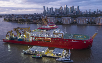 A large red ship is floating in a river with a city skyline behind it.